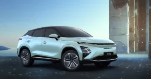 A light blue 2025 Chery OMODA E5 electric SUV is parked on a dark, reflective surface. The car is angled towards the viewer, with its front left wheel prominently displayed. The background features a futuristic, abstract architectural structure on the right and a serene, watery landscape with a hint of a distant landmass on the left