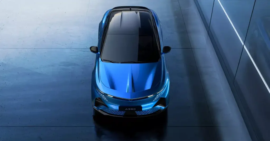 A top-down, high-angle shot showcases a sleek, futuristic blue 2026 Alpine A390 car with a black panoramic roof. The car is positioned centrally on a polished, dark gray floor, reflecting the ambient blue and white lighting. The background features a modern architectural element with clean lines and subtle vertical lighting strips, creating a sense of depth and sophistication.