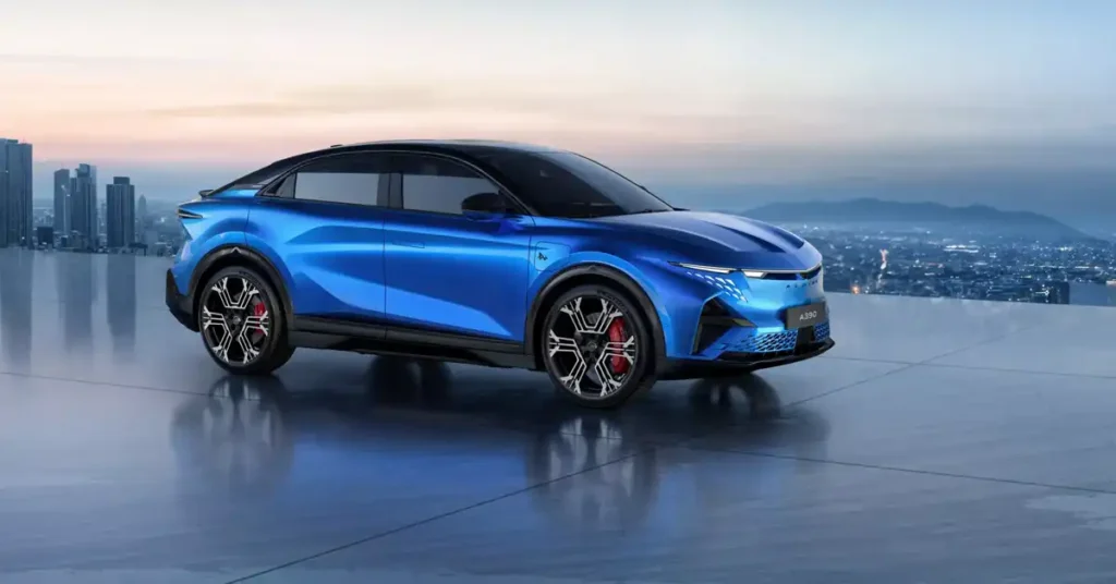 A sleek, blue electric 2026 Alpine A390SUV is positioned on a ref lective surface, with a cityscape and mountains in the background under a soft, twilight sky. The car is angled towards the viewer, showcasing its aerodynamic design, sharp headlights, and distinctive wheels with red brake calipers.