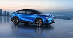 A sleek, blue electric 2026 Alpine A390SUV is positioned on a ref lective surface, with a cityscape and mountains in the background under a soft, twilight sky. The car is angled towards the viewer, showcasing its aerodynamic design, sharp headlights, and distinctive wheels with red brake calipers.