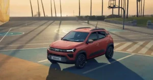 A bright orange 2026 Dacia Spring electric car is parked on a basketball court at sunset. The car is positioned in the center of the frame, angled slightly towards the viewer. The basketball court has blue and orange sections, with white lines marking the boundaries. In the background, palm trees cast long shadows across the court, and a lifeguard tower and a beach can be seen in the distance.