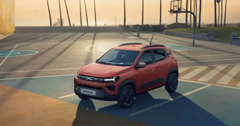 A bright orange 2026 Dacia Spring electric car is parked on a basketball court at sunset. The car is positioned in the center of the frame, angled slightly towards the viewer. The basketball court has blue and orange sections, with white lines marking the boundaries. In the background, palm trees cast long shadows across the court, and a lifeguard tower and a beach can be seen in the distance.