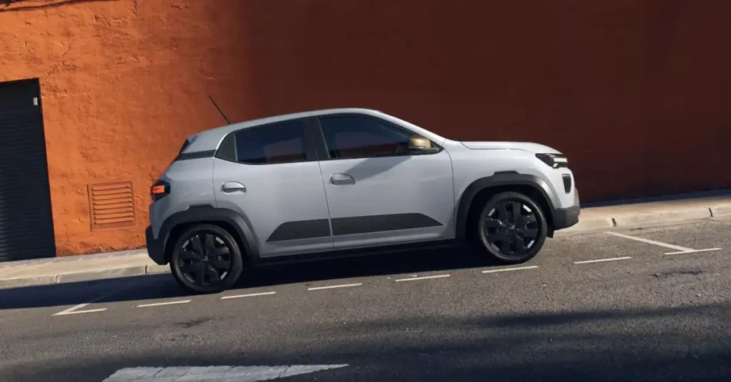 A light gray, 2026 Dacia Spring electric car is parked on an asphalt road with white parking lines. The car is viewed from the side, with its front facing right. It has black trim around the wheel wells and black rims. Behind the car is a sidewalk and a textured, orange-brown wall with a dark gray door and a ventilation grate. The lighting suggests it's daytime, with shadows cast on the road.