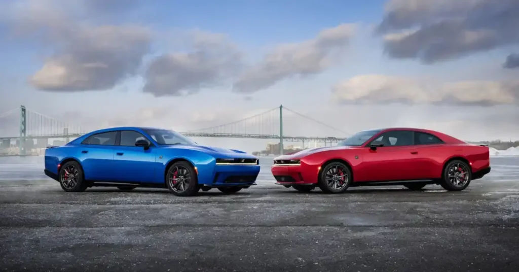 A blue four-door 2026 Dodge Charger Daytona sedan and a red two-door coupe are parked facing each other on a wet, asphalt surface. Both cars are modern muscle cars with aggressive styling, featuring sharp lines and prominent grilles.