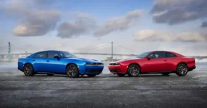 A blue four-door 2026 Dodge Charger Daytona sedan and a red two-door coupe are parked facing each other on a wet, asphalt surface. Both cars are modern muscle cars with aggressive styling, featuring sharp lines and prominent grilles.