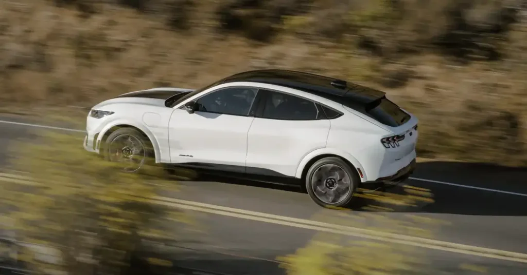 A white Ford Mustang Mach-E with black racing stripes is driving on a winding road along a coastal cliffside. The sun is setting, casting a warm glow over the scene. The car is in motion, with its wheels blurred to indicate speed. The background features rolling hills and the ocean.