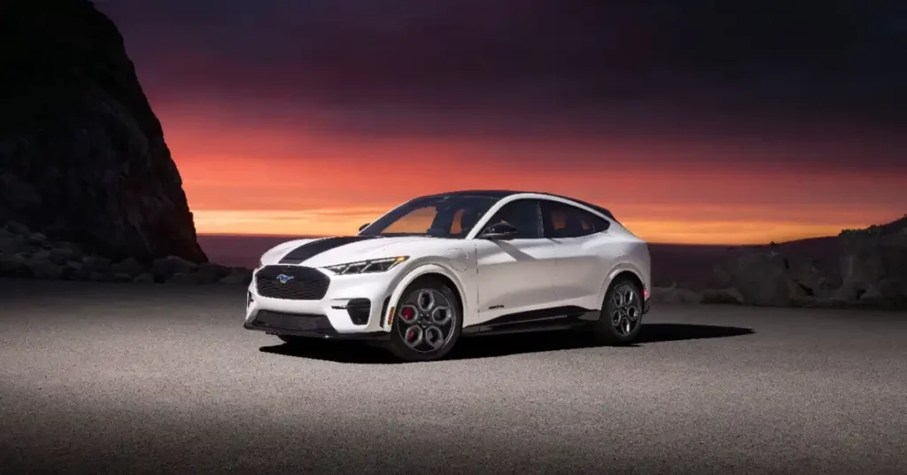 A white2026 Ford Mustang Mach-E GT is parked on a paved surface. The car has black racing stripes on the hood and a black roof. The background features a dramatic sunset with vibrant orange and purple hues.