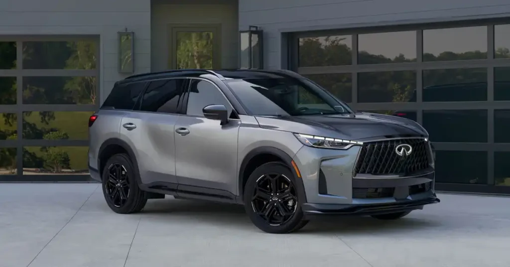 A silver 2026 INFINITI QX60 with a black roof and hood is parked in front of a modern garage with glass doors. The SUV is angled towards the viewer, showcasing its sleek design and black rims.