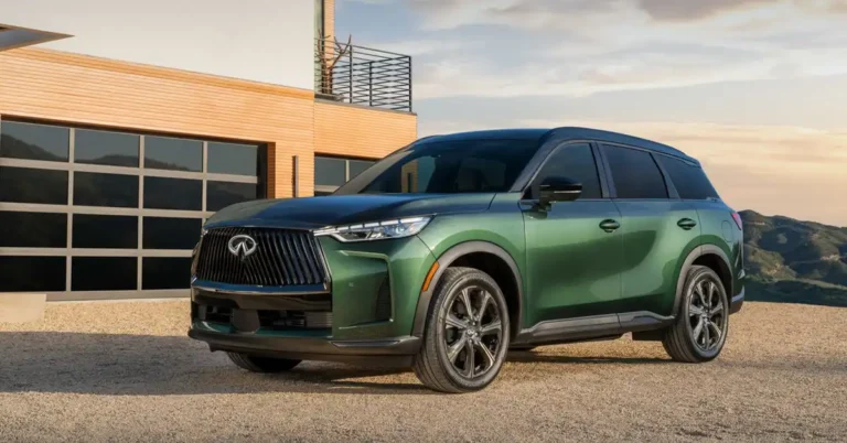 A dark green 2026 INFINITI QX60 SUV is parked on a gravel driveway in front of a modern house with wood paneling and a large glass garage door. The SUV is angled towards the viewer, with its front grille and headlights clearly visible. The background shows rolling hills under a soft, hazy sky, suggesting a sunset or sunrise.