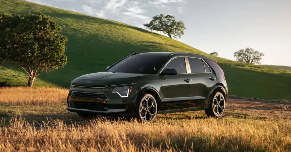 A dark green 2026 Kia Niro Hybrid is parked in a field of dry grass, with a rolling green hill in the background. Two trees are visible on the hill, one larger and closer to the car, and another smaller one further away.