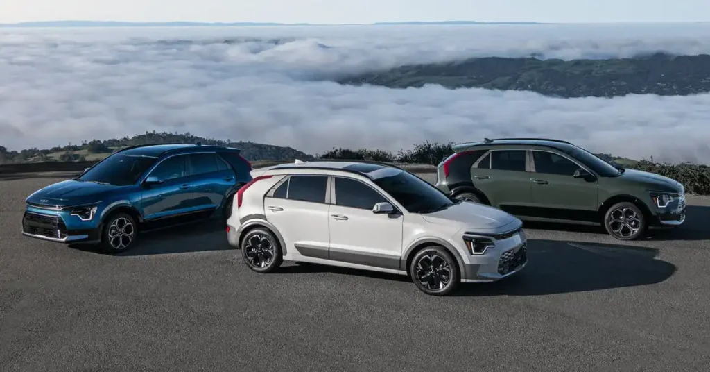 Three 2026 Kia Niro Hybrid SUVs are parked on a paved surface with a dramatic backdrop of clouds and rolling hills. The car in the foreground is white, the one to its left is blue, and the one to its right is green.