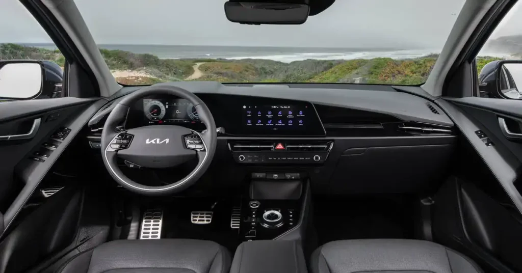 A wide-angle interior shot of a 2026 Kia Niro Hybrid SUV dashboard, featuring a sleek black interior. The steering wheel, with the Kia logo prominently displayed, is on the left. To its right, a large, integrated touchscreen displays various icons and the time.