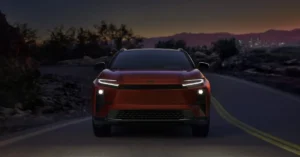 A front-facing shot of a red 2026 Toyota C-HR EV SUV driving on a road at dusk. The SUV's headlights are on, illuminating the road ahead. The background features a dark, mountainous landscape with a distant city skyline.