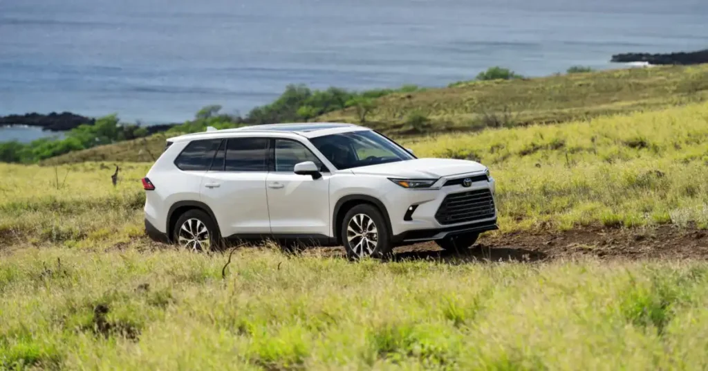 A white 2026 Toyota Grand Highlander is parked on a dirt road in a grassy field. The SUV is facing to the right, with its front end slightly angled towards the camera. The background shows a body of water, possibly the ocean, with some rocky islands and green hills.