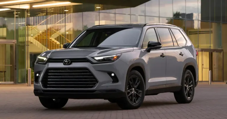 A gray 2026 Toyota Grand Highlander is parked in front of a modern building with large glass windows and an illuminated staircase visible inside. The SUV is angled towards the camera, showcasing its front grille, headlights, and black alloy wheels.