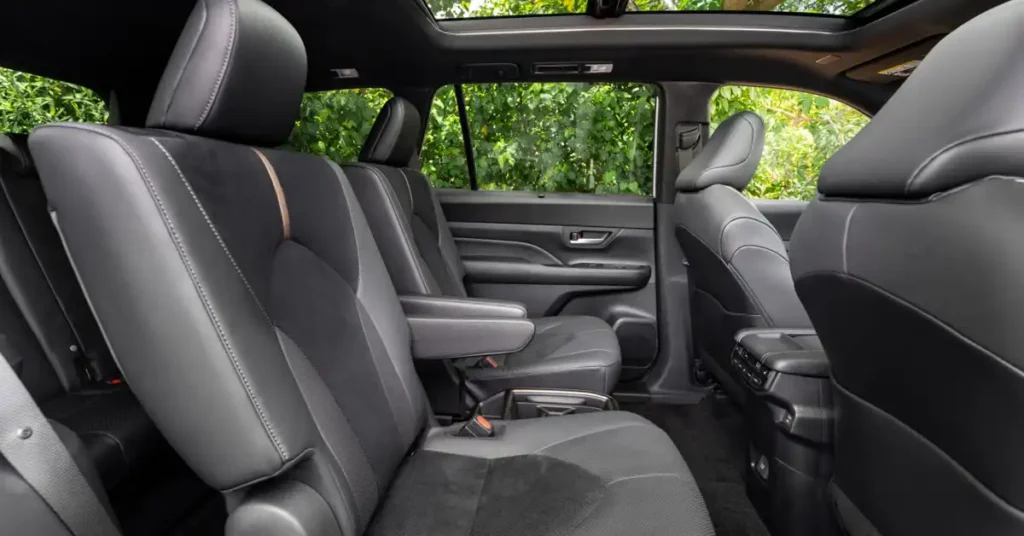 A wide shot captures the luxurious interior of a 2026 Toyota Grand Highlander, focusing on the rear seating area. The seats are upholstered in a combination of black leather and a textured dark fabric, with subtle stitching and a thin, elegant copper accent on the headrest of the first row.