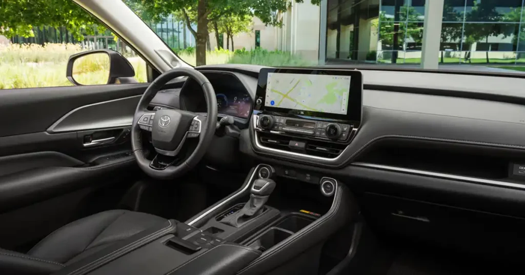 The image shows the interior of a 2026 Toyota Grand Highlander, specifically the driver's side dashboard and center console. The focus is on the sleek, dark interior with a large touchscreen display showing a navigation map.