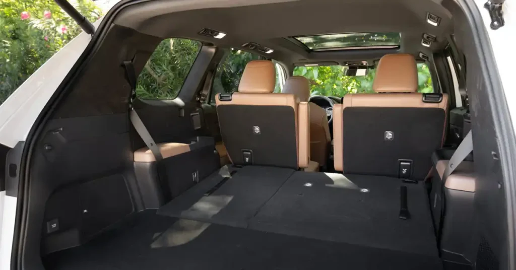 The image shows the interior of a white 2026 Toyota Grand Highlander with the rear hatch open, revealing the cargo area and the back of the second-row seats. The seats are upholstered in tan leather with black fabric backing, and they are folded down to create a flat cargo space.