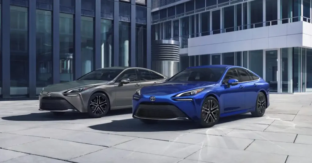 Two 2026 Toyota Mirai sedans, one blue and one silver, are parked on a paved outdoor area in front of a modern building with large glass windows. The blue car is in the foreground, angled towards the viewer, while the silver car is slightly behind and to the left. The building's architecture is sleek and contemporary, with clean lines and reflective surfaces.