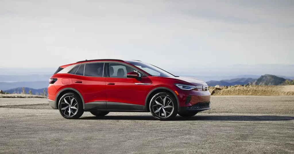 A red Volkswagen ID.4 electric SUV is parked on a paved road with a mountainous landscape in the background. The car is viewed from the side, with its front facing right. The sky is clear and bright, with a hint of haze in the distance.