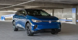 A blue Volkswagen ID.4 electric SUV is parked in a concrete parking garage. The car is facing the camera at a slight angle, with its headlights on. The background shows the textured concrete ceiling and walls of the garage, with some blue barriers and railings visible in the distance.