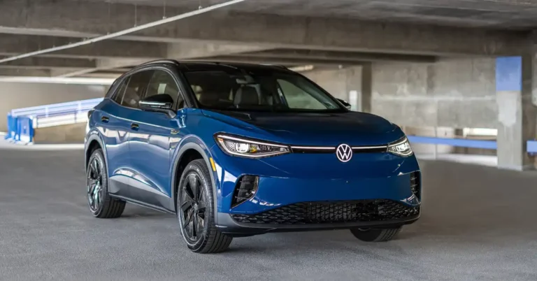 A blue Volkswagen ID.4 electric SUV is parked in a concrete parking garage. The car is facing the camera at a slight angle, with its headlights on. The background shows the textured concrete ceiling and walls of the garage, with some blue barriers and railings visible in the distance.