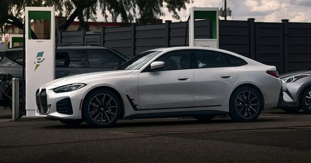 A white BMW i4 is parked at an electric vehicle charging station. The car is positioned in the center of the frame, viewed from the side. To the left of the BMW, an Electrify America charging station is visible, with its distinctive green and white branding.