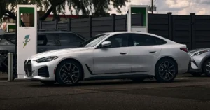 A white BMW i4 is parked at an electric vehicle charging station. The car is positioned in the center of the frame, viewed from the side. To the left of the BMW, an Electrify America charging station is visible, with its distinctive green and white branding.