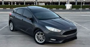 dark gray Ford Focus hatchback is parked in an outdoor parking lot. The car is angled towards the viewer, with its front left wheel closest to the camera. The car has tinted windows and a sunroof. In the background, there is a row of green bushes, a road with white lines, and a modern building with a light-colored facade and dark windows.