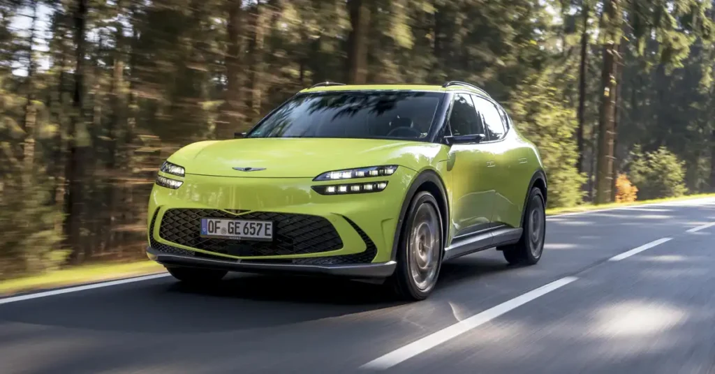 A bright green Genesis GV60 electric SUV is captured in a dynamic, low-angle shot as it drives on a paved road through a forest.