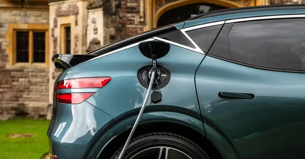 A close-up, side-view shot focuses on the rear quarter panel of a teal Genesis GV60 Electric car as it is being charged. The charging port is open, with a gray charging cable plugged in. The car's sleek design is highlighted by its glossy finish, which reflects the surrounding environment.