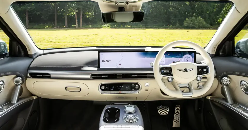 The image shows the interior of a Genesis GV60 Electric car viewed from the driver's perspective. The dashboard is dominated by a large, wide infotainment screen displaying a map and the time. To the right of the screen is a white steering wheel with integrated controls.