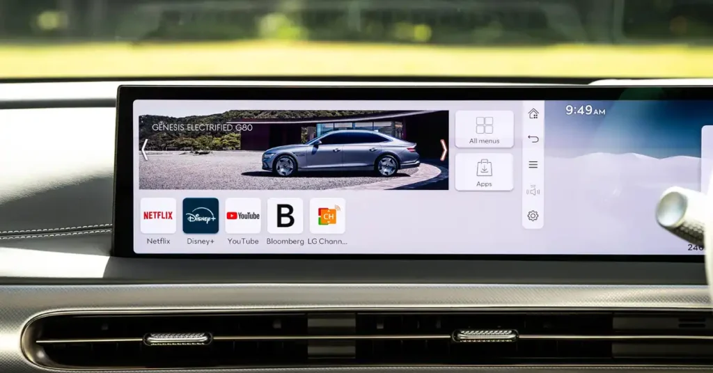 A wide shot shows a car's infotainment screen displaying a Genesis Electrified G60. The screen is divided into sections: the top shows the car with a modern building in the background, and the bottom displays app icons for Netflix, Disney+, YouTube, Bloomberg, and LG Channels. To the right of the car image, there are icons for "All menus" and "Apps." The time "9:49 AM" is visible in the top right corner.