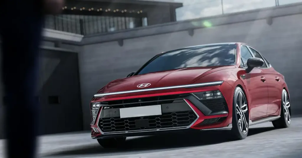 A red Hyundai Sonata N Line sedan is parked in an urban environment. The car is angled towards the viewer, with its front end prominently displayed. The background features a modern building with a gray concrete facade and a railing with lights. The car's design is aggressive, with sharp lines, a distinctive front grille, and sleek LED headlights.