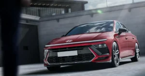 A red Hyundai Sonata N Line sedan is parked in an urban environment. The car is angled towards the viewer, with its front end prominently displayed. The background features a modern building with a gray concrete facade and a railing with lights. The car's design is aggressive, with sharp lines, a distinctive front grille, and sleek LED headlights.