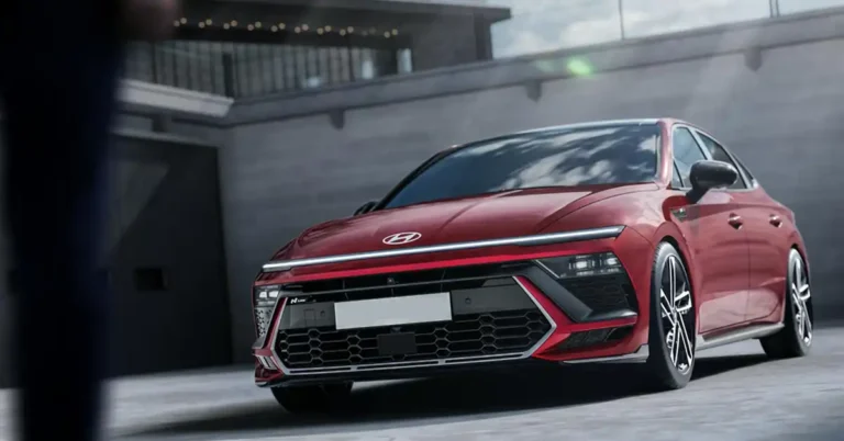 A red Hyundai Sonata N Line sedan is parked in an urban environment. The car is angled towards the viewer, with its front end prominently displayed. The background features a modern building with a gray concrete facade and a railing with lights. The car's design is aggressive, with sharp lines, a distinctive front grille, and sleek LED headlights.