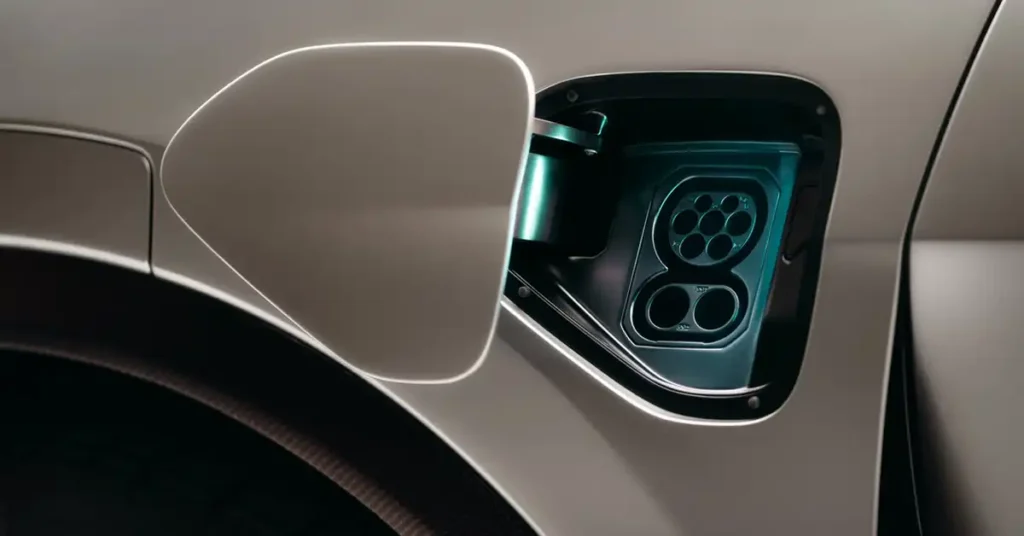 A close-up, low-angle shot focuses on the open charging port of a Lotus Eletre electric SUV. The port, with its intricate array of circular connectors, is illuminated by a cool, teal light, highlighting its futuristic design.