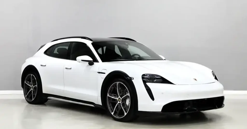 A white Porsche Taycan Cross Turismo is parked in a studio with a light gray background. The car is angled towards the viewer, with its front left side most prominent. It has a black roof and black wheel arches, contrasting with its white body. The car's headlights are illuminated, and its wheels are a dark gray with silver accents.