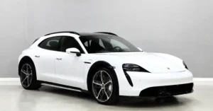 A white Porsche Taycan Cross Turismo is parked in a studio with a light gray background. The car is angled towards the viewer, with its front left side most prominent. It has a black roof and black wheel arches, contrasting with its white body. The car's headlights are illuminated, and its wheels are a dark gray with silver accents.