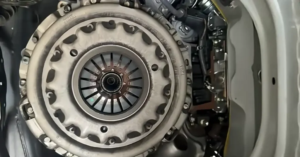 A close-up, overhead shot focuses on a car's clutch assembly. The central element is the clutch pressure plate, a large, circular metal component with a serrated outer edge and numerous bolts securing it.