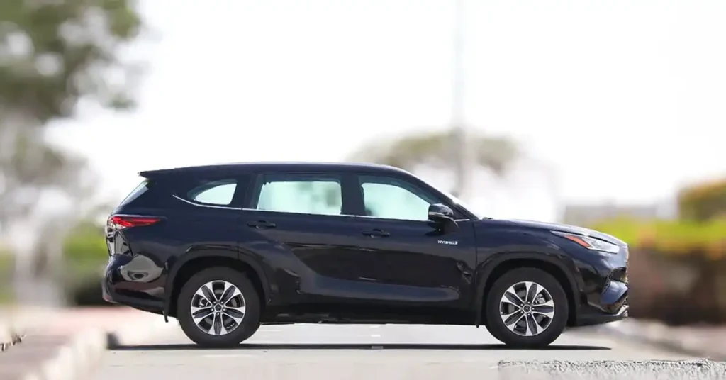A dark blue Toyota Highlander Hybrid SUV is shown in a side profile view, parked on a paved surface. The car is the main subject, with its sleek design and alloy wheels clearly visible.