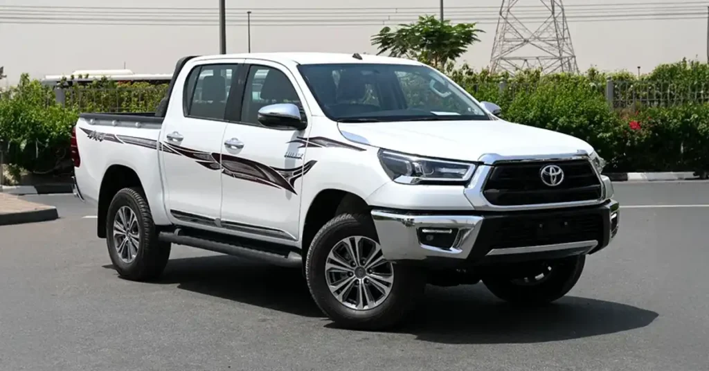 A white Toyota Hilux pickup truck is parked on an asphalt road. The truck is facing the viewer at a slight angle, with its front left side most prominent. It has chrome accents on the grille, bumper, and side mirrors, and a dark graphic decal runs along the side of the truck bed and doors.