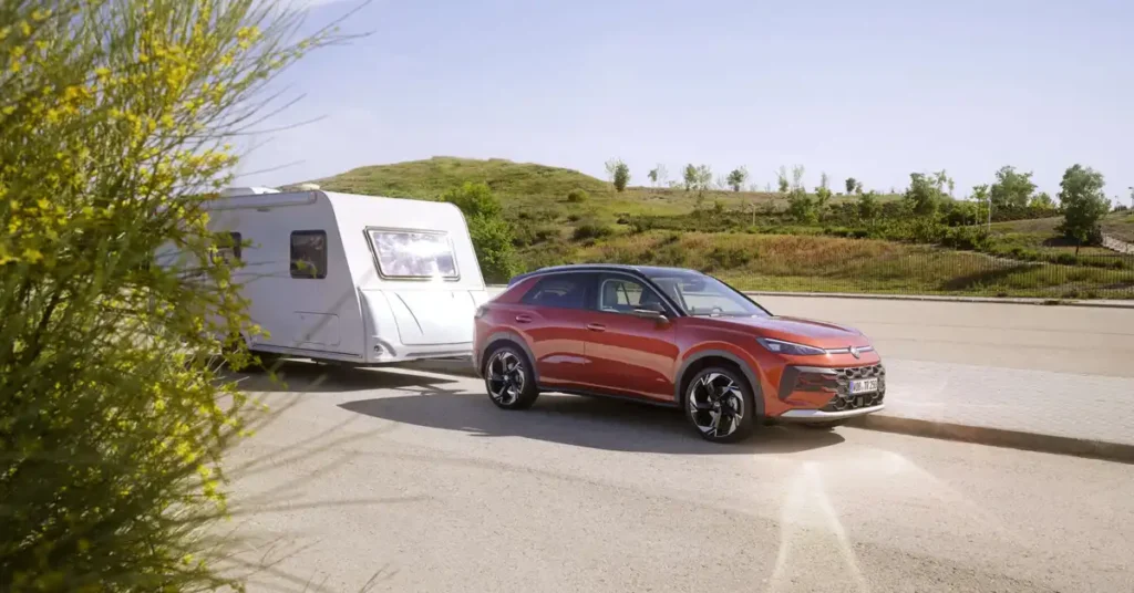 A bright, sunny day with a clear blue sky. In the foreground, a vibrant orange Volkswagen T-Roc 2026 is hitched to a white caravan, parked on a paved surface. The SUV's wheels are turned slightly, suggesting it's either parked or maneuvering.