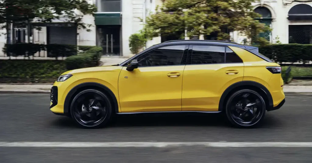 A bright yellow VVolkswagen T-Roc 2026 is captured in a side profile, moving from left to right on a paved road. The car features a black roof and black wheel arches, with black alloy wheels that show motion blur.