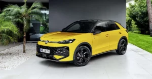 A bright yellow Volkswagen T-Roc 2026 is parked on a light-colored tiled driveway. The car has a black roof and black rims. To the left of the car, there is a palm tree with lush green fronds, and a pile of white gravel. Behind the car, a modern building with dark gray walls is visible, and to the right, a well-maintained green lawn and trees create a natural backdrop.