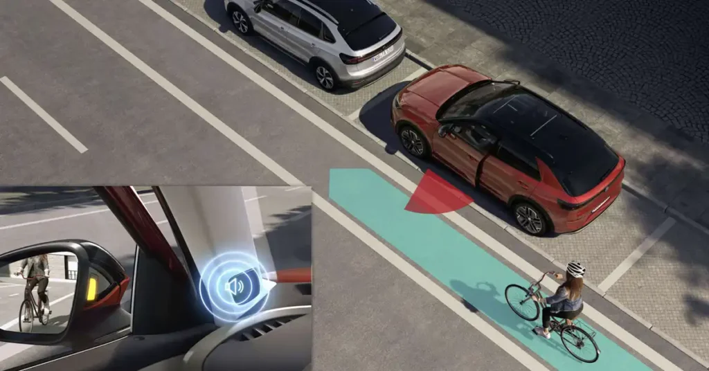 An aerial view shows a redVolkswagen T-Roc 2026 with its door open, parked next to a silver car on a street. A cyclist is riding on a designated bike lane next to the red car. A graphic overlay highlights the car's blind-spot monitoring system, showing a red warning zone and a cyclist in the side mirror. The scene is set on a sunny day with shadows cast on the road.