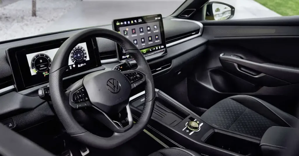 A close-up, eye-level shot showcases the interior of aVolkswagen T-Roc 2026, focusing on the driver's side. The prominent, perforated leather steering wheel with the Volkswagen logo is centered in the foreground. To the left, a digital dashboard displays driving information, including speed and navigation. To the right, a large touchscreen infotainment system is visible, displaying various app icons.
