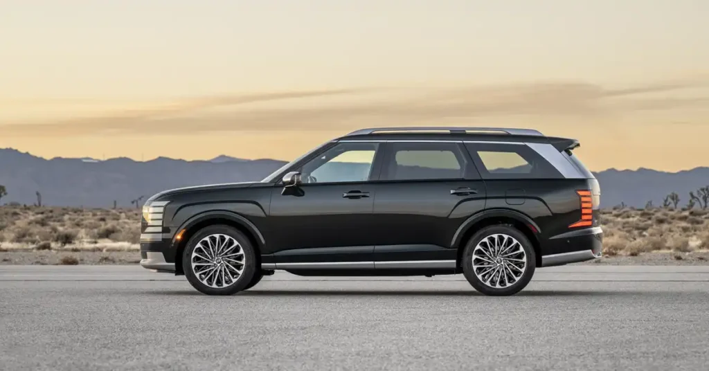 A dark gray 2026 Hyundai Palisade is parked on an asphalt road in a desert landscape. The SUV is shown in profile, with the front facing left.