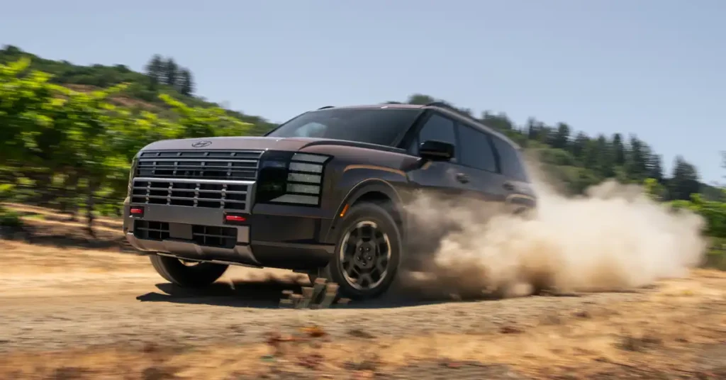 A brown 2026 Hyundai Palisade SUV is captured mid-drift on a dirt road, kicking up a large cloud of dust. The vehicle is angled towards the viewer, with its front left wheel turned sharply.