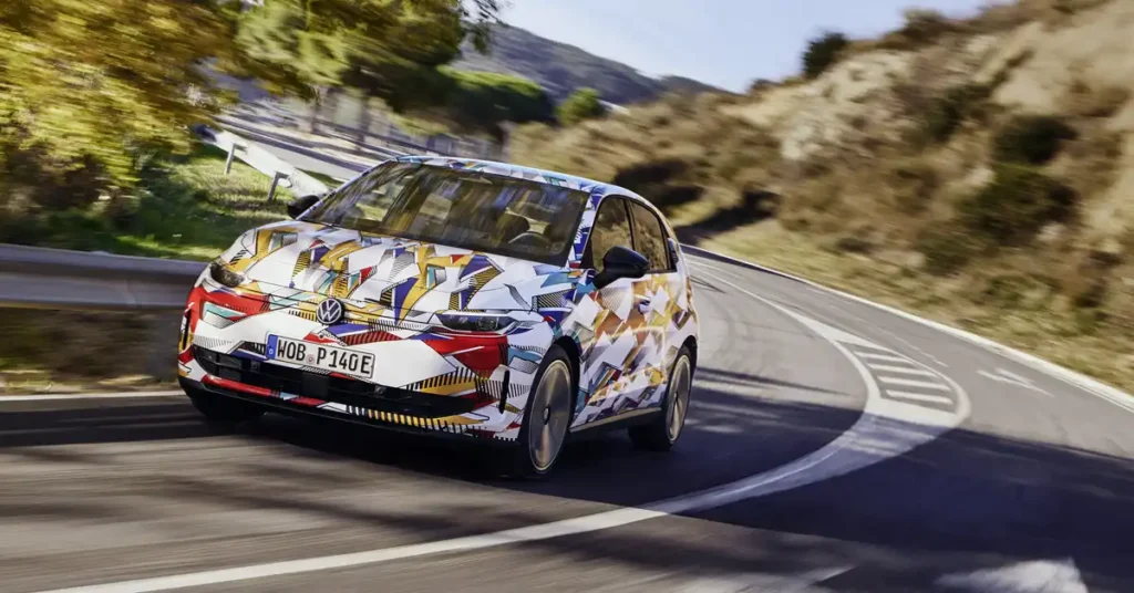 A 2026 Volkswagen ID. Polo electric car with a colorful, abstract geometric camouflage wrap is driving on a winding road. The car is angled towards the viewer, with its front left wheel slightly turned. The road has white lane markings and is surrounded by dry, hilly terrain with sparse vegetation.